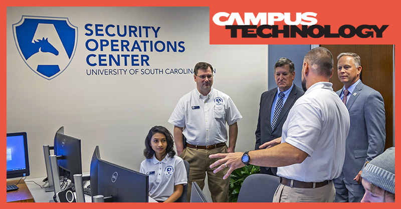Five adults stand and sit talking in a room of computers and the sign on the wall says "Security Operations Center University of South Carolina Aiken"